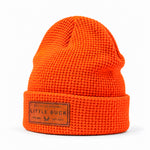 Bright orange waffle-knit beanie hat with a folded cuff and a brown rectangular leather patch reading LITTLE BUCK on a white background.